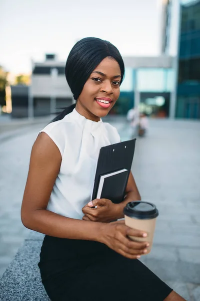 Business woman with cardboard coffee cup and notepad resting during lunch break outdoors, office building on background. Black businesswoman in skirt and white blouse