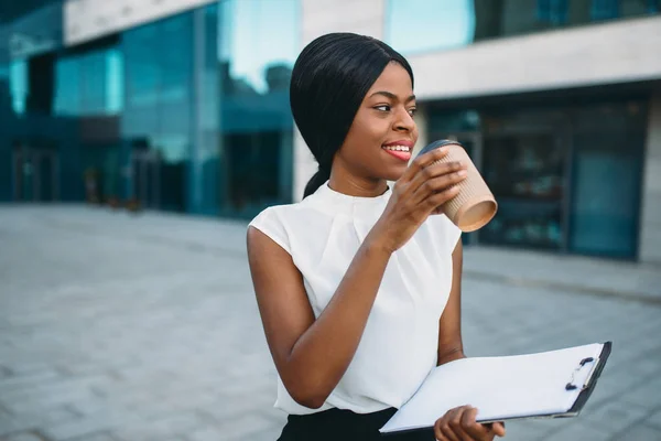 Black business girl grinks coffee from cardboard cup outdoors, office building on background. Black businesswoman in white blouse on working break