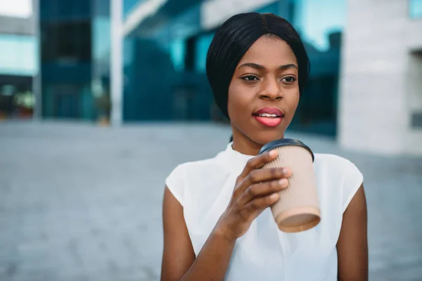 Black business girl grinks coffee from cardboard cup outdoors, office building on background. Black businesswoman in white blouse on working break