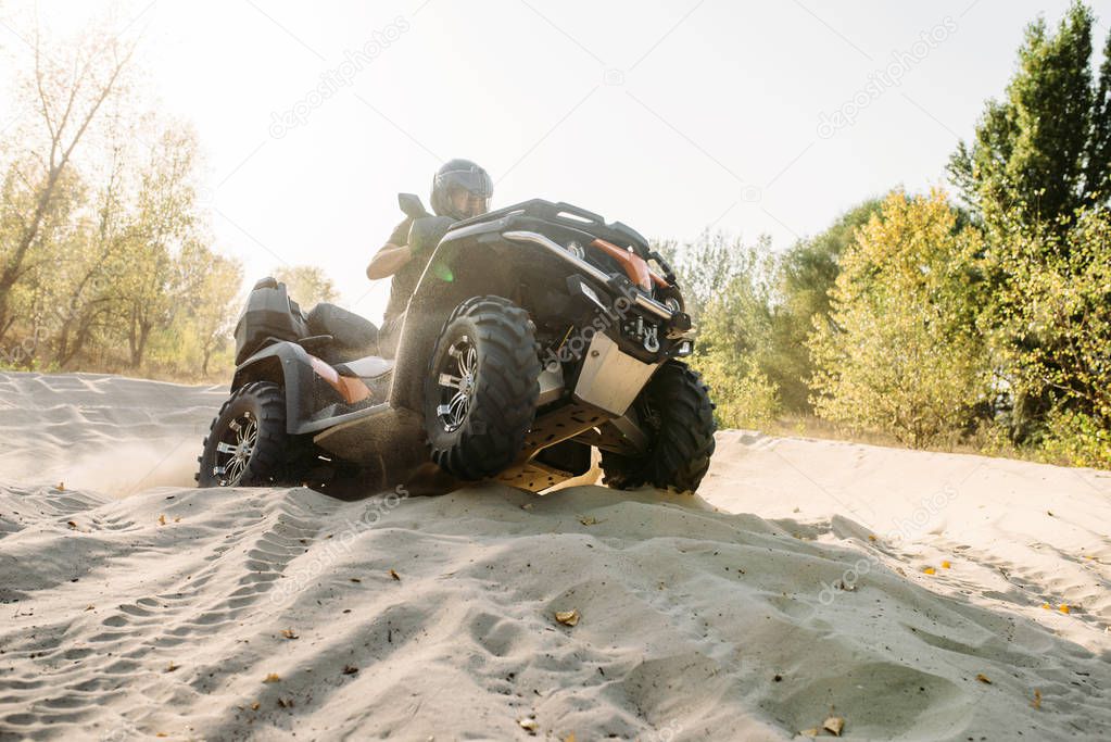 Atv rider in helmet climbing sandy road in forest, sunshine on background. Offroad riding on quad bike, extreme sport and travelling, quadbike adventure