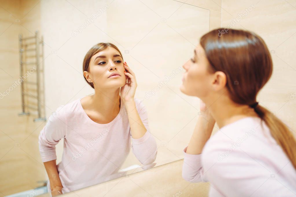 Young woman looking on face at the mirror in bathroom. Female person cares for skin. Morning facial cleaning procedure
