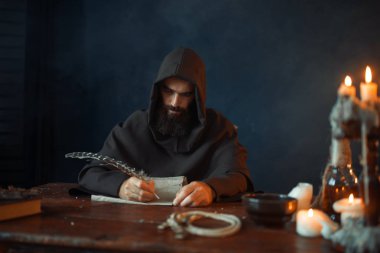 Ortaçağ keşiş masada oturan ve yazar bir kaz tüyü, üstten görünüm, gizli din ile. Karanlık pelerinli gizemli rahip. Gizem ve Maneviyat