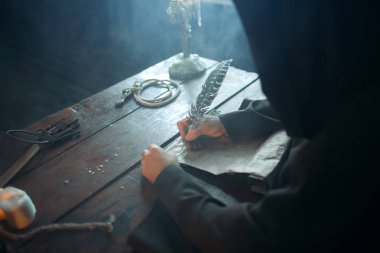 Ortaçağ keşiş masada oturan ve yazar bir kaz tüyü, üstten görünüm, gizli din ile. Karanlık pelerinli gizemli rahip. Gizem ve Maneviyat