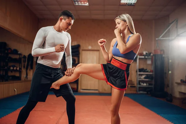 Female person makes a kick in the groin, self defense workout with male personal trainer, gym interior on background. Woman on training, self-defense practice