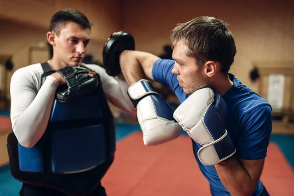 Male kickboxer in gloves practicing elbow kick with a personal trainer in pads, workout in gym. Fighter doing a powerful punch on training, kickboxing practice in action