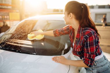Young woman with sponge scrubbing vehicle glass with foam, car wash. Lady on self-service automobile washing. Outdoor carwash at summer day  clipart