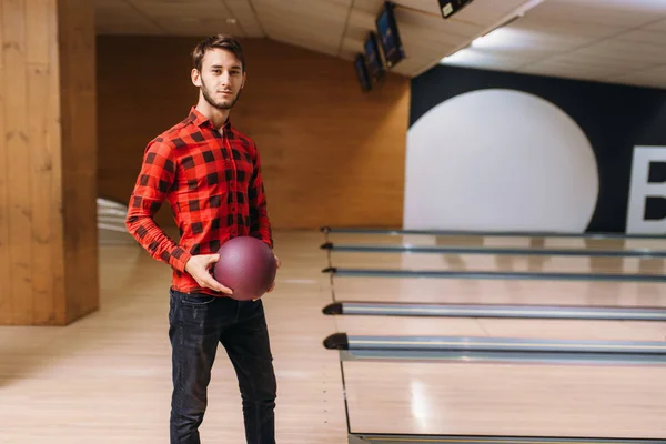 Male bowler standing on lane and holds ball in hands, back view. Bowling alley player poses in club, active leisure