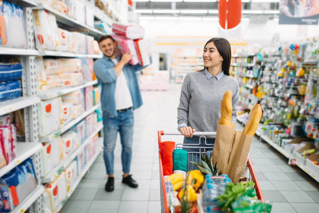 Happy couple with cart buying a lot of diapers in a supermarket, family shopping. Customers in shop, buyers in market, department of goods for children