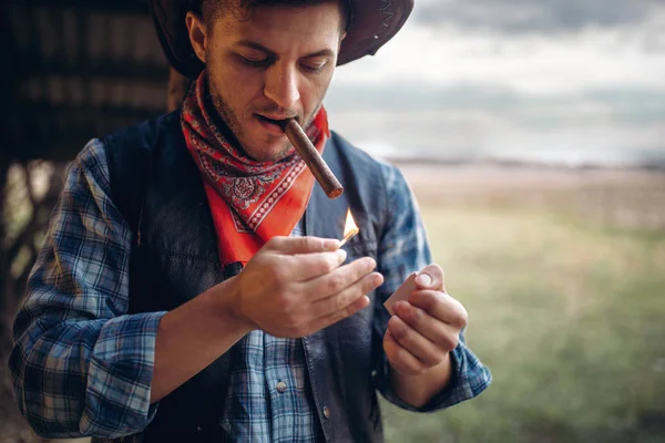 Bearded cowboy lights a cigar with matches, texas ranch on background, western. Vintage male person in leather clothes on farm, wild west culture