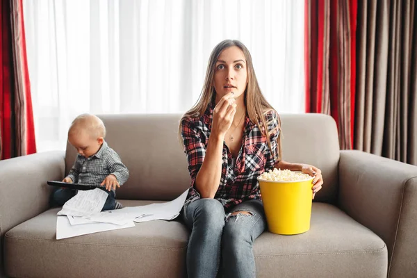 Mother watch tv and eats popcorn, little male kid plays near. Mom and son happy together at home, togetherness