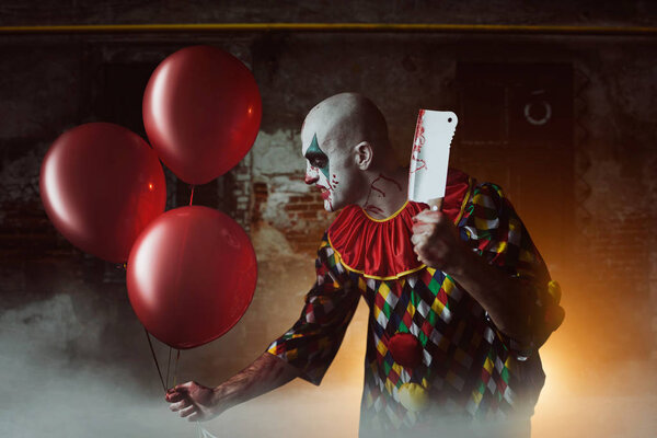 Scary bloody clown with meat cleaver and air balloons sneaking into the basement, horror. Man with makeup in carnival costume, crazy maniac