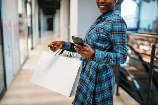 Black female person with phone and shopping bags in mall. Shopaholic in clothing store, consumerism lifestyle, fashion