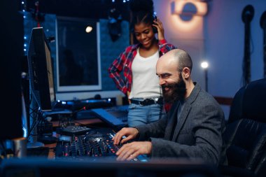 Sound engineer and female singer at remote control panel in audio recording studio. Musician in headphones listens composition, professional music mixing clipart