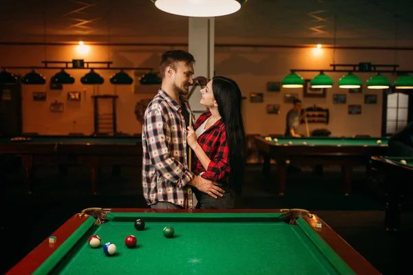 Couple hugs at the table with colorful balls in billiard room. Man and woman plays american pool game in sport bar