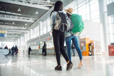 Uluslararası Havaalanının bekleme alanında sırt çantaları ile iki kadın turist. Hava terminali, arka görünüm, beyaz ve siyah bayanlar mutlu yolculuk, mutlu kadınların yaz seyahat bagaj ile yolcular