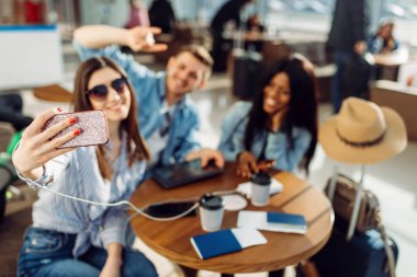 Üç turist havaalanında telefonda selfie yapar. Bagaj ile yolcu hava terminalinde uçuş için sabırsızlanıyoruz, seyahat mutlu arkadaşlar, tatile yaz seyahat