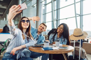 Üç turist havaalanında telefonda selfie yapar. Bagaj ile yolcu hava terminalinde uçuş için sabırsızlanıyoruz, seyahat mutlu arkadaşlar, tatile yaz seyahat