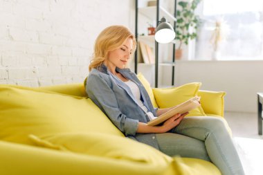 Genç kadın rahat sarı kanepede bir kitap okuma, arka planda beyaz tonlarda oturma odası. Kanepe üzerinde oturan dergi ile çekici kadın kişi