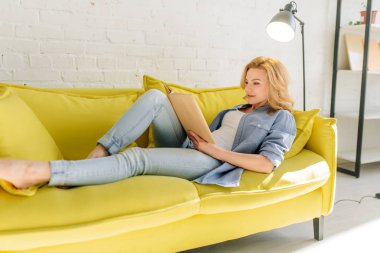 Young woman lying on cozy yellow couch and reading a book, living room in white tones on background. Attractive female person with magazine sitting on sofa at home