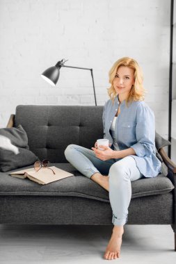 Kitap ve kahve fincan ile genç bayan rahat siyah kanepede pozlar, arka planda beyaz tonlarda oturma odası. Evde kanepede oturan dergi ile çekici kadın kişi