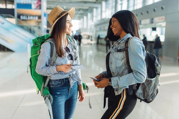 国際空港でバックパックを持つ2人の女性観光客 エアターミナルで手荷物をお持ちのお客様 バックビュー 白と黒の女性の幸せな旅 幸せな女性の夏の旅 — ストック写真
