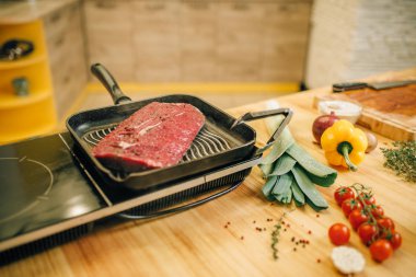 Elektrikli sobanın üzerinde tavada kavrulmuş et bonfile, arka planda tahta da sebzeler, kimse. Tezgahüzerinde otlar ile Sığır eti, baharat ve baharatlar