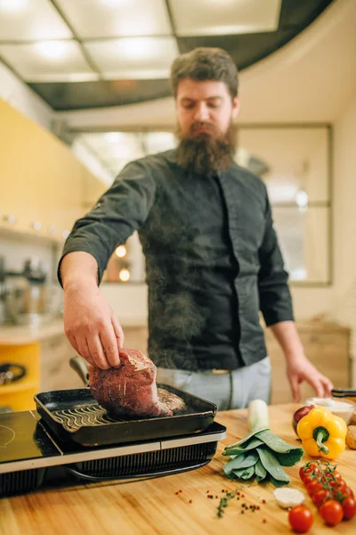 ひげを生やしたシェフが台所のフライパンで肉を調理 テーブルの電気ストーブの上でゆでた豚肉を準備する男 — ストック写真