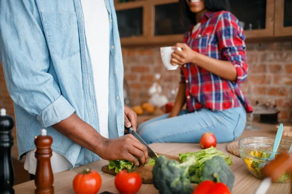 man cooking breakfast on the kitchen, wife drinks coffee. African couple preparing vegetable salad at home. Healthy vegetarian lifestyle