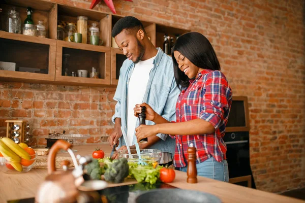 Black man cooking breakfast on the kitchen, wife drinks coffee. African couple preparing vegetable salad at home. Healthy vegetarian lifestyle