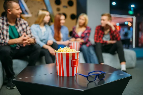 Group of teenagers relax on the couch and waiting for showtime in cinema hall. Male and female youth sitting on sofa in movie theater, popcorn on the table