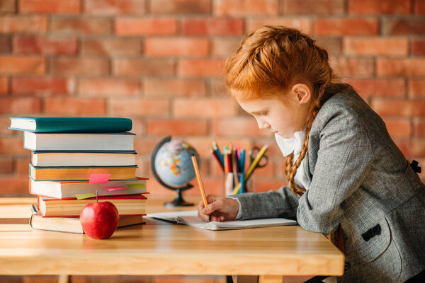 Cute schoolgirl doing homework at the table with textbooks, apples and globe. Female pupil reading a book at the desk