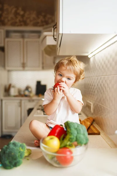 テーブルの上に座ってトマトを食べる小さな女の子 女性の赤ちゃんは台所のボウルから野菜を味わいます 子供はベジタリアン料理を味わう — ストック写真