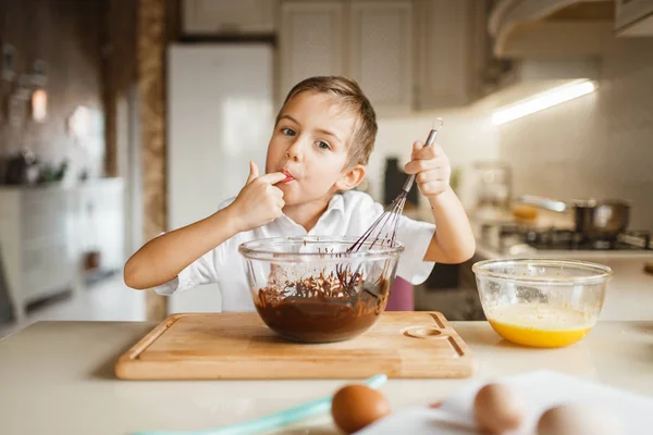 Male kid tastes melted chocolate in a bowl, pastry preparation. Cute boy cooking on the kitchen. Happy child prepares sweet dessert at the counter