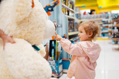 Little girl choosing big teddy bear in kids store, side view. Daughter looking for toys in supermarket, family shopping, young customer clipart
