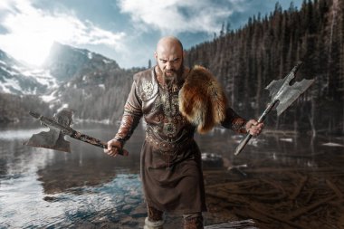 Ellerindeki baltalarla birlikte, gölde duran geleneksel Nordic kıyafetleri giymiş Viking. Nehirde eski savaşçı, arka planda kayalık dağlar