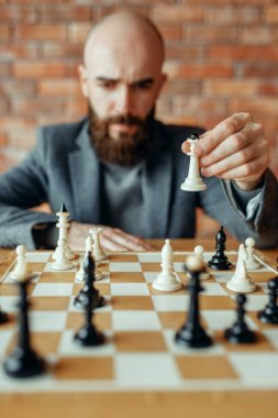 Erkek satranç oyuncusu beyaz figürler oynarken, kraliçe hareket. Satranç oyuncusu, ön görüş, entelektüel turnuva kapalı. Ahşap masa üzerinde Satranç Tahtası, strateji oyunu