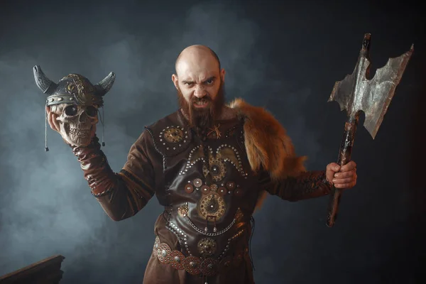 伝統的な北欧の服を着た怒っているバイキングは ヘルメットと斧 野蛮なイメージ 殺人者で敵の頭蓋骨を保持しています 煙の中の古代の戦士 — ストック写真