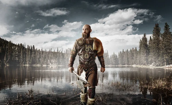 身着传统北欧服装的手持斧头的帅气 站在湖边 古代战士在河 北森林的背景 — 图库照片