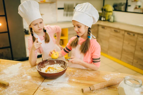 Two little girls cooks in caps rubs lemon to the bowl, cookies preparation on the kitchen. Kids cooking pastry, children chefs preparing cake