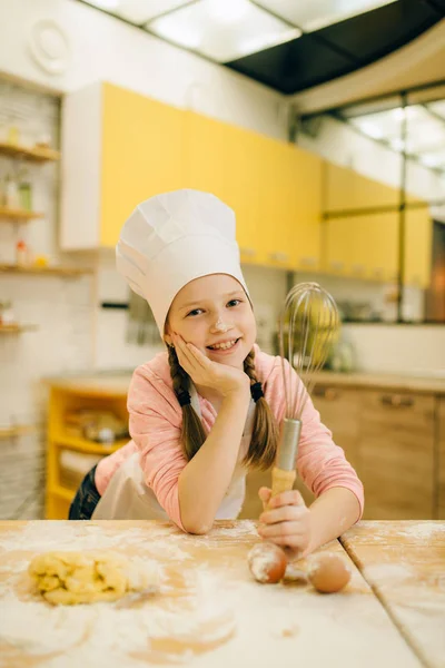 キャップとエプロンで小さな女の子の料理は キッチンで混合 クッキーの準備のための泡立て器を保持しています 子供たちはペストリーを調理し ケーキを準備する子供のシェフ — ストック写真