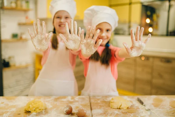 キャップで2人の小さな女の子が調理し 小麦粉で覆われた手 キッチンでクッキーの準備を示しています 子供たちはペストリーを調理し ケーキを準備する子供のシェフ — ストック写真