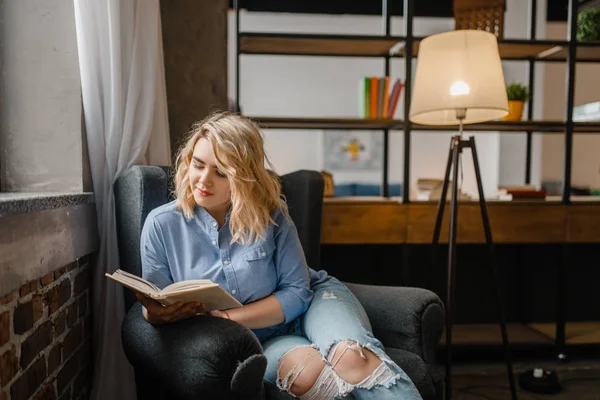椅子に座って本を読んでいる若い女性 女性は快適なソファ 背景にリビングルームのインテリアで読みます — ストック写真