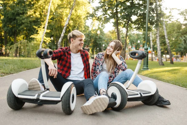 年轻的夫妇坐在马路上与陀螺板在夏季公园 带电动陀螺板的户外休闲活动 生态运输 电动陀螺仪车辆 — 图库照片