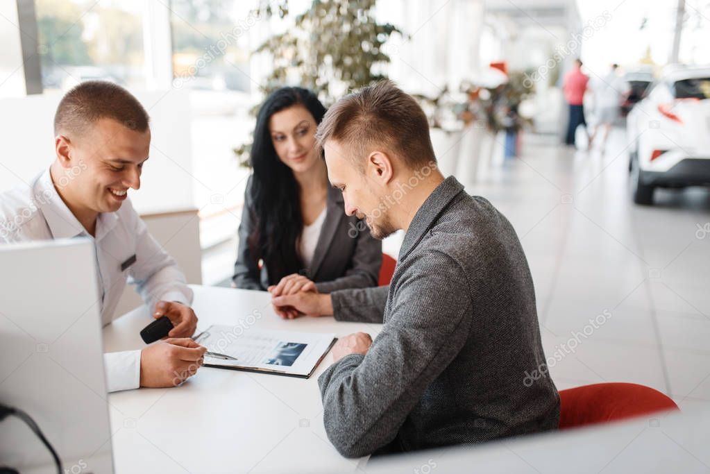 Salesman and a couple make out the sale of a new car in the showroom. Male and female customers looks vehicle in dealership, automobile sale, auto purchase