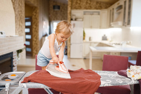 Little baby irons clothes on the kitchen. Kid doing housework at home. Young housewife clean the house