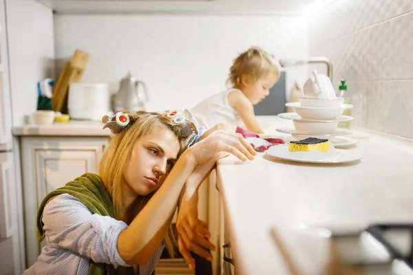 疲れた主婦の近くで皿洗いをする小さな女の子 家で家事をしている赤ちゃんを持つ女性 彼女の娘と一緒に女性は家をきれいに — ストック写真