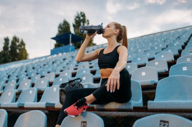 Tribünde oturan ve su içen, stadyumda antrenman yapan kadın bir koşucu. Kadın dışarıda koşmadan önce esneme egzersizi yapıyor.