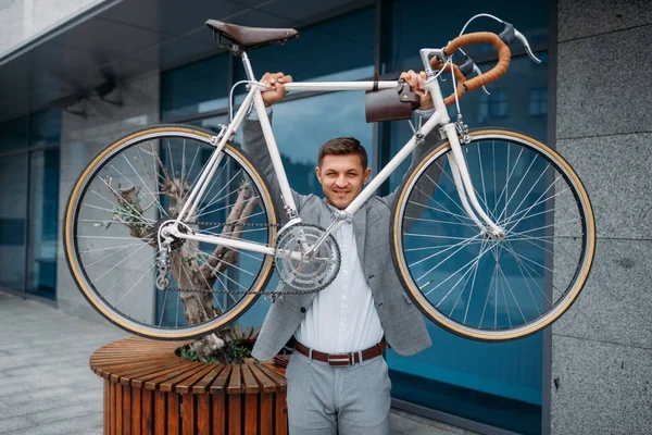 Strong businessman holds bicycle at the glass office building in downtown. Business person riding on eco transport on city street, urban style