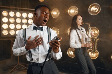Sahnede sahne ışıklarıyla saksafoncusu olan erkek jazzman ve kadın saksafoncu. Sahnede çalan caz sanatçıları
