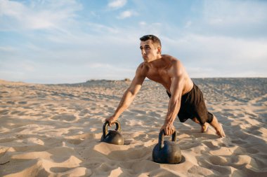Erkek sporcu, güneşli bir günde çölde iki tane kettlebell ile şınav egzersizi yapıyor. Sporda güçlü motivasyon, açık hava eğitiminde güçlü.